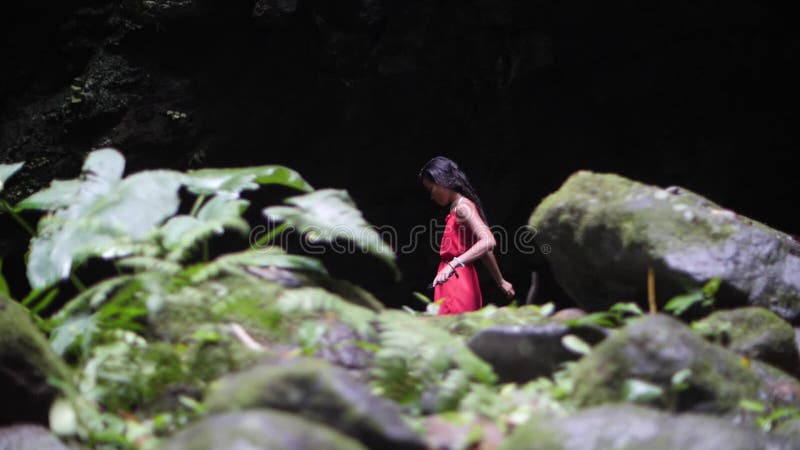 Joven vestida de rojo caminando sola cerca de una oscura cueva en la jungla.