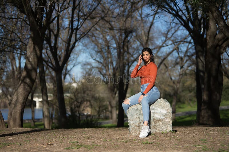 Joven y bonita mujer con camiseta naranja y jeans mirando la cámara relajada y tranquila sentada sobre una enorme piedra en un par imagen de archivo libre de regalías