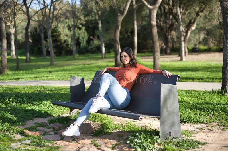 Joven y bonita mujer de camisa naranja y jeans mirando la cámara relajada y tranquila sentada en un banco de metal en un parque. 4 foto de archivo