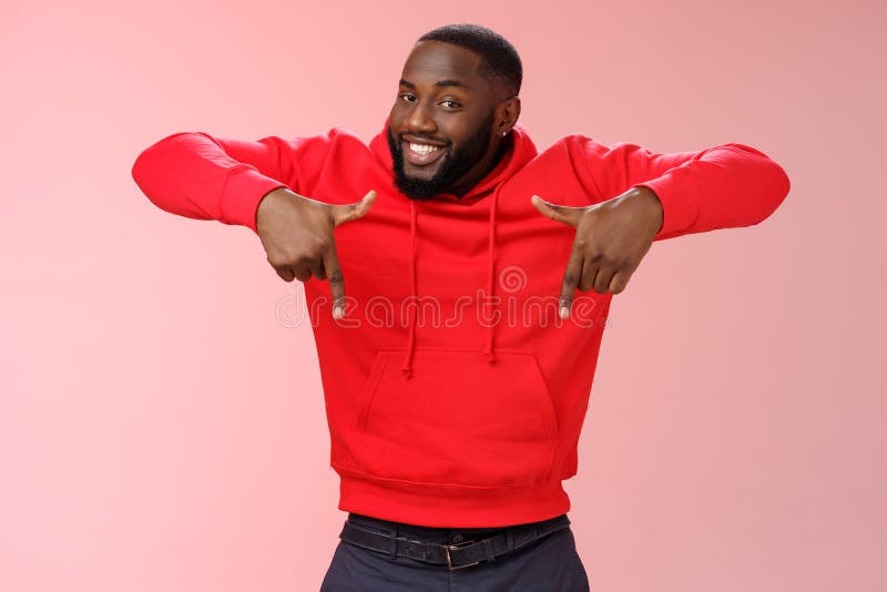 Joven y carismático hombre negro barbudo y apuesto, con capucha roja, apuntando hacia abajo los dedos índice sonriendo mostrando a imágenes de archivo libres de regalías