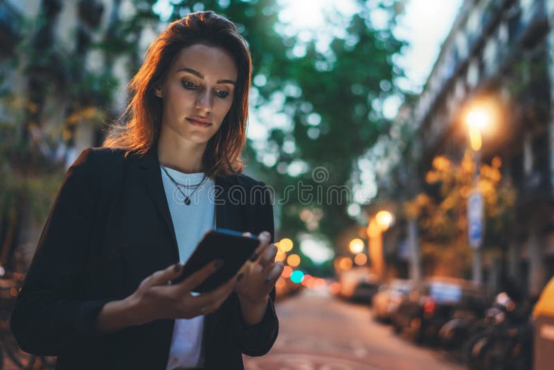 Young stylish business woman writes message on smartphone walking evening city street, hipster tourist traveling in european town and planning trip using mobile phone. Young stylish business woman writes message on smartphone walking evening city street, hipster tourist traveling in european town and planning trip using mobile phone