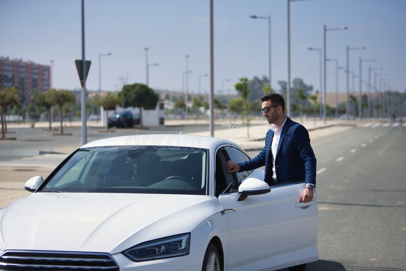 Joven y exitoso hombre hispano con chaqueta y gafas de sol abriendo la puerta de su auto de lujo blanco. coches de concepto foto de archivo libre de regalías