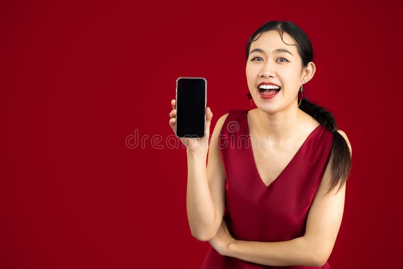 Young beautiful asian woman wear elegant red dress holding and showing blank screen smartphone on red color background. Young beautiful asian woman wear elegant red dress holding and showing blank screen smartphone on red color background