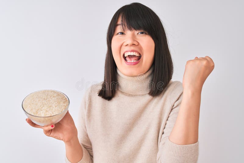 Joven y hermosa mujer china sosteniendo el tazón con arroz sobre fondo blanco aislado gritando orgullosa y celebrando la victoria imagen de archivo