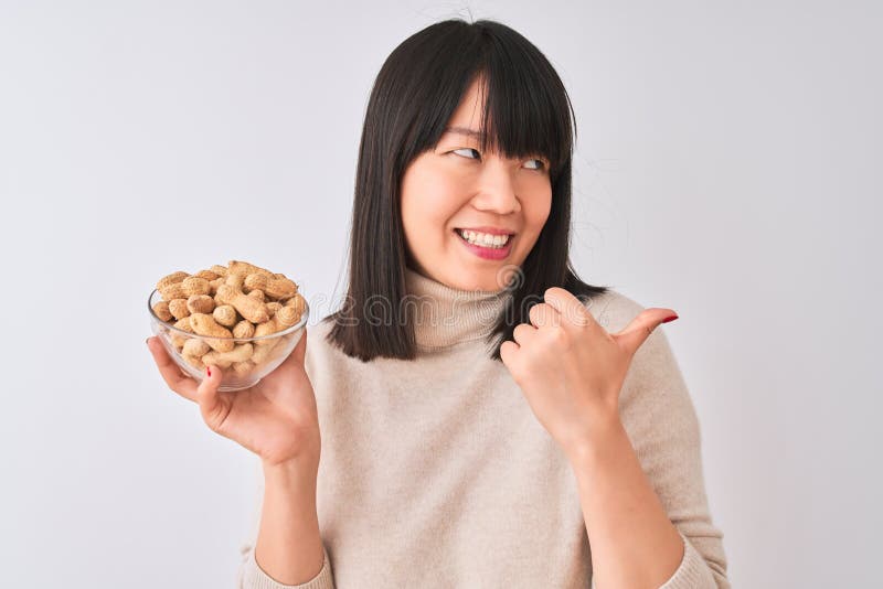 Joven y hermosa mujer china sosteniendo un tazón con cacahuetes sobre fondo blanco aislado apuntando y mostrando con pulgar hacia foto de archivo libre de regalías