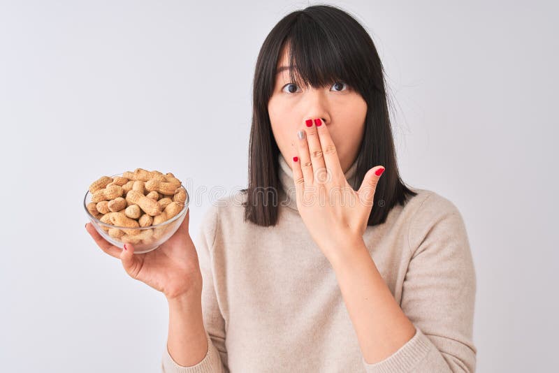Joven y hermosa mujer china sosteniendo un tazón con cacahuetes sobre fondo blanco aislado boca cubierta con la mano impactada con imagen de archivo libre de regalías