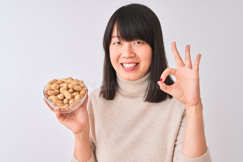 Joven y hermosa mujer china sosteniendo un tazón con cacahuetes sobre fondo blanco aislado haciendo buen signo con los dedos excel fotos de archivo