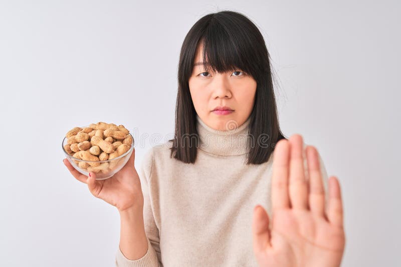 Joven y hermosa mujer china sosteniendo un tazón con cacahuetes sobre fondo blanco aislado con la mano abierta haciendo signo de d imagenes de archivo