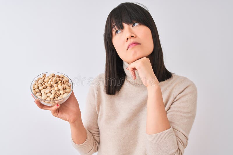 Joven y hermosa mujer china sosteniendo un tazón con pistachos sobre fondo blanco aislado cara seria pensando en la pregunta foto de archivo libre de regalías