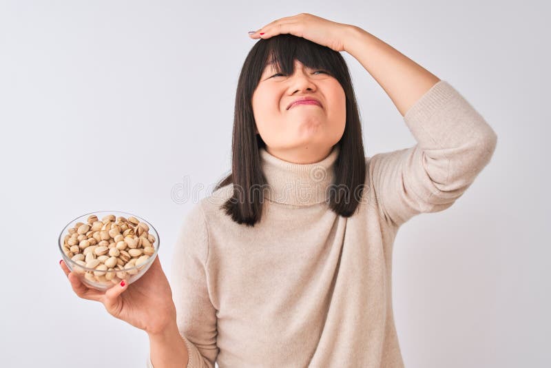 Joven y hermosa mujer china sosteniendo un tazón con pistachos sobre fondo blanco aislado estresado con la mano en la cabeza impac foto de archivo