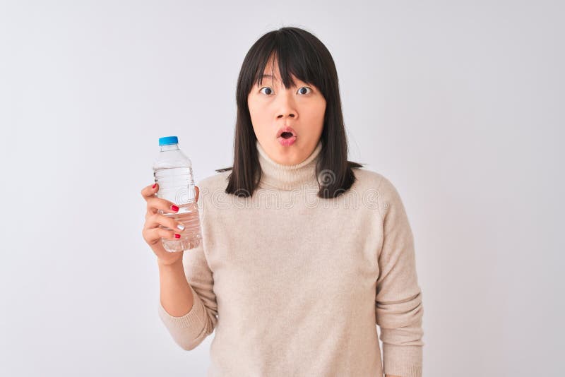 Joven y hermosa mujer china sostiene una botella de agua sobre fondo blanco aislado asustada en shock con una cara sorpresa asusta imagen de archivo