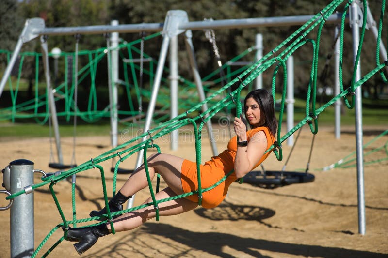 Joven y hermosa mujer con un vestido naranja mirando la cámara relajada y tranquila tendida en una red de cuerda en un parque. bel imágenes de archivo libres de regalías