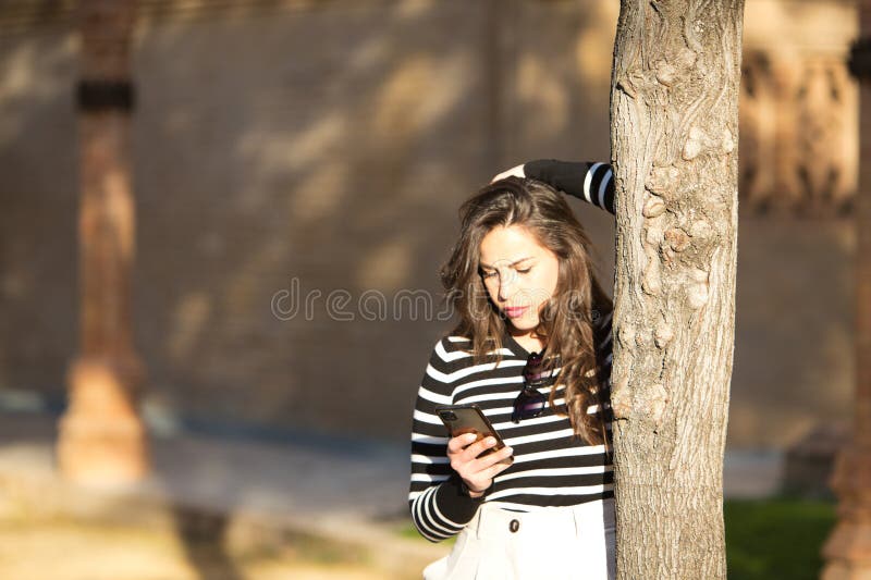 Joven y hermosa mujer de largo pelo castaño con una camiseta de rayas blancas y negras apoyada en un árbol mirando su móvil fotografía de archivo