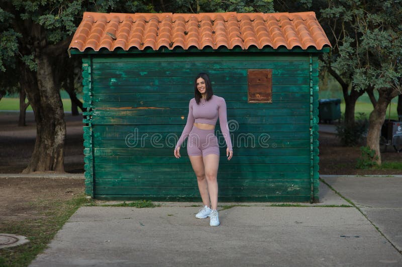 Joven y hermosa mujer deportista vestida de deporte caminando feliz y sonriendo con cabaña de madera verde al fondo. deporte conce foto de archivo