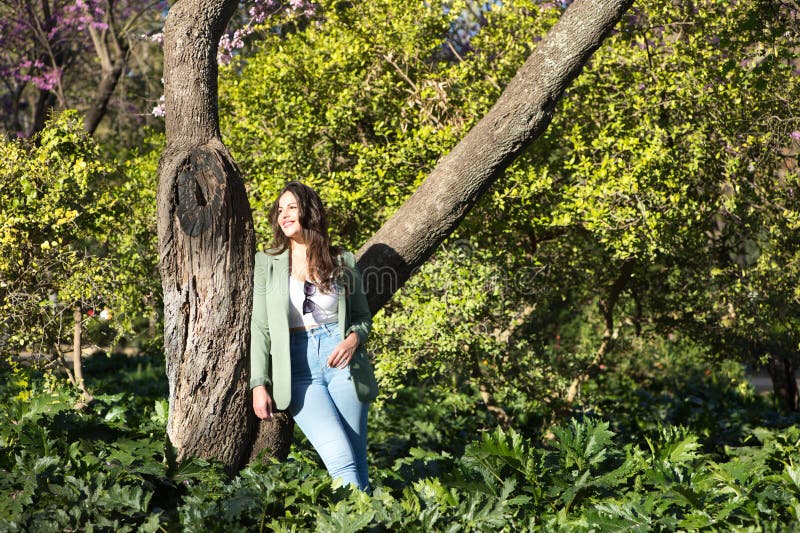 Joven y hermosa mujer española de cabello largo y marrón vestido con jeans y chaqueta verde apoyándose en un árbol en el parque de foto de archivo libre de regalías