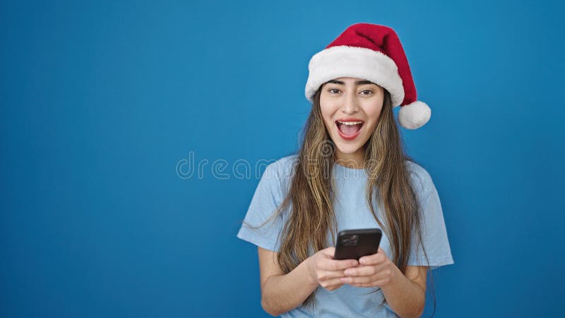 Joven y hermosa mujer hispana con sombrero de navidad usando smartphone sobre fondo azul aislado fotos de archivo