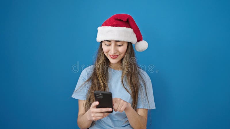 Joven y hermosa mujer hispana con sombrero de navidad usando smartphone sobre fondo azul aislado imagen de archivo