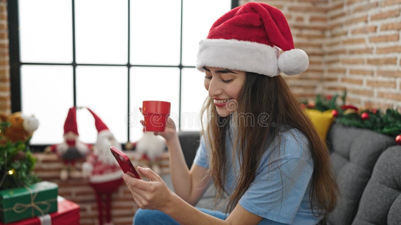 Joven y hermosa mujer hispana usando un smartphone tomando café celebrando navidades en casa foto de archivo