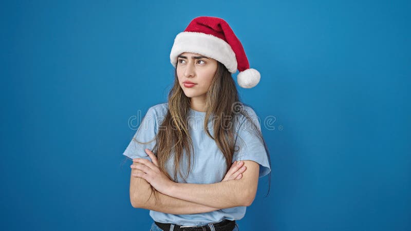 Joven y hermosa mujer hispánica de pie disgustada usando sombrero de navidad sobre fondo azul aislado foto de archivo libre de regalías