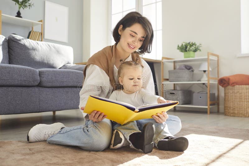 Joven y sonriente madre sentada en el piso con un bebé que lee un libro de cuento de hadas juntos en casa. imágenes de archivo libres de regalías