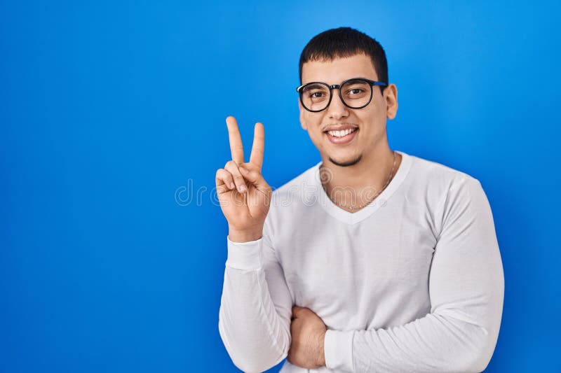 Joven árabe con camisa blanca casual y gafas sonriendo con cara feliz guiñando el ojo en la cámara haciendo signo de victoria imagen de archivo