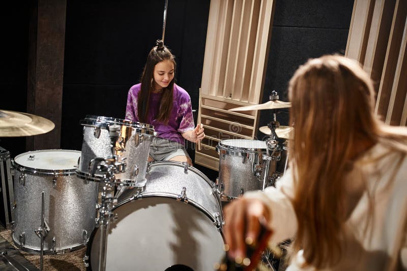 jolly adorable teenage girl playing drums while her blonde friend looking at her, musical group, stock photo. jolly adorable teenage girl playing drums while her blonde friend looking at her, musical group, stock photo