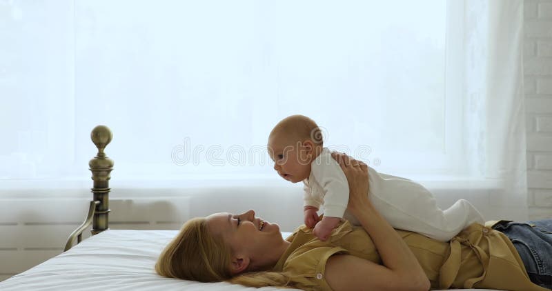 Jovencita alegre tendida en la cama levantando a su adorable bebé