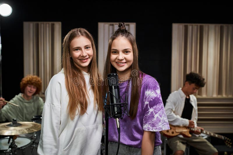 jolly teenage girls looking at camera near microphone while their friends playing drums and guitar, stock photo. jolly teenage girls looking at camera near microphone while their friends playing drums and guitar, stock photo