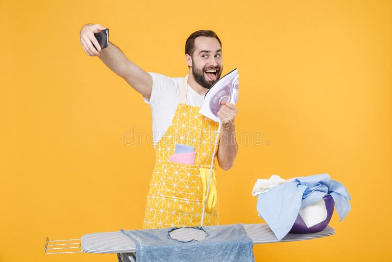 Jovencito gracioso esposo de la casa planchando ropa limpia a bordo haciendo tareas domésticas aisladas de fondo amarillo fotografía de archivo