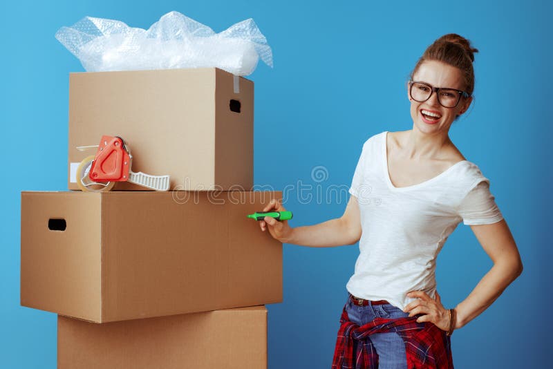 Happy young woman in white t-shirt signs cardboard box isolated on blue. woman labeling cardboard box with green color permanent marker on side of a box. Happy young woman in white t-shirt signs cardboard box isolated on blue. woman labeling cardboard box with green color permanent marker on side of a box