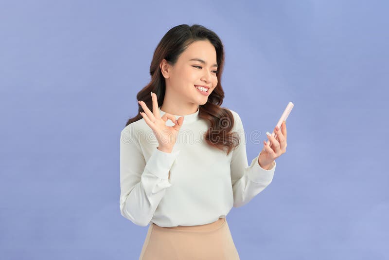 Joyeuse asiatique femme tenant smartphone et montre ok signe sur fond violet clair photographie stock libre de droits