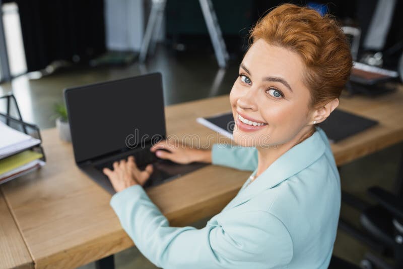 Joyeuse femme d'affaires rousse tapant sur floue. Joyeuse femme d'affaires rousse dactylographiant sur ordinateur portable floue avec écran blanc et souriant à l'appareil photo dans l'image du stock de bureau