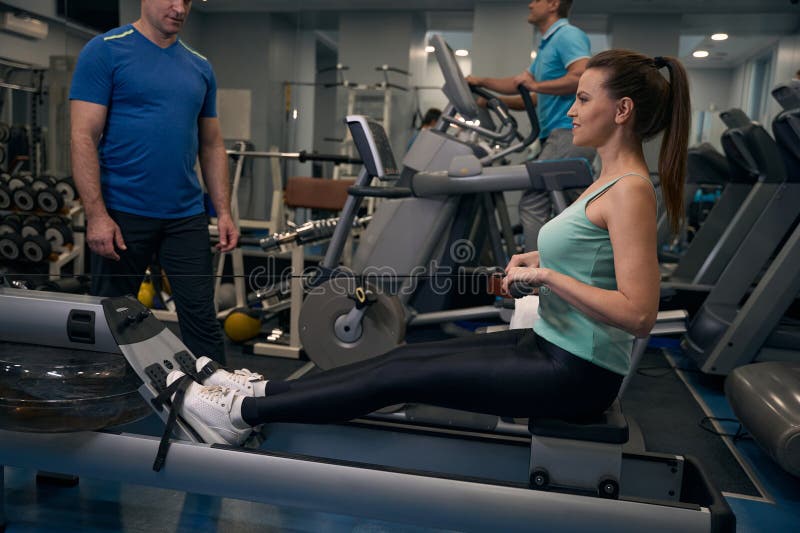 Joyeuse femme s'entraîner sur des équipements de gym supervisés par un entraîneur image libre de droits