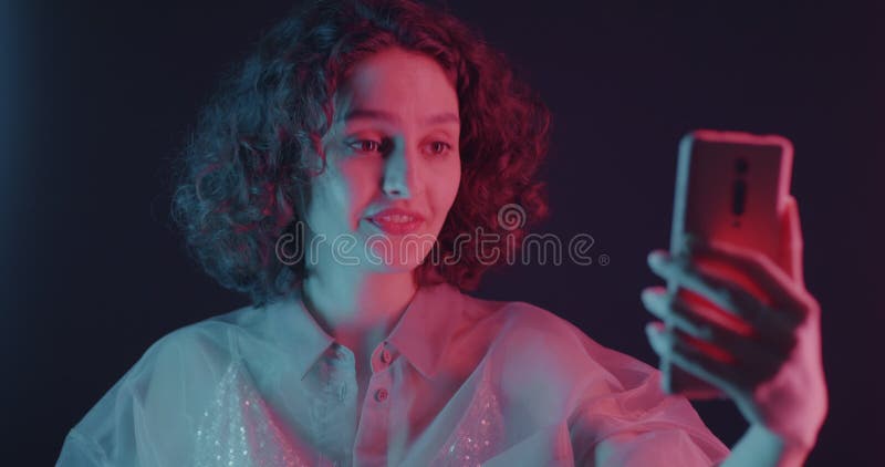 Joyeuse jeune femme faisant appel vidéo en ligne avec smartphone dansant en néon léger banque de vidéos