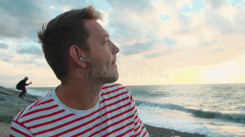 Joyeux jeune homme caucasien écoutant de la musique avec des écouteurs utilisant un smartphone sur la côte de la mer et profitant banque de vidéos