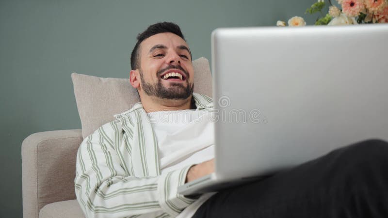 Joyeux jeune homme riant avec coeur en utilisant un ordinateur portable sur un canapé confortable en profitant d'un délicieux mome banque de vidéos