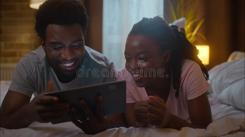 Joyeux rires heureux famille afro-américaine couple ethnique femme homme allongé lit regarder la tablette numérique soutien en lig banque de vidéos