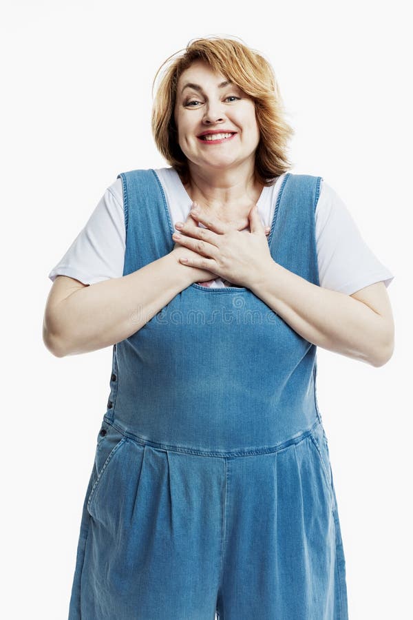 A joyful beautiful middle-aged woman in denim overalls and a white T-shirt emotionally pressed her hands to her chest. Activity stock photo A joyful beautiful middle-aged woman in denim overalls and a white T-shirt emotionally pressed her hands to her chest. Activity stock photo