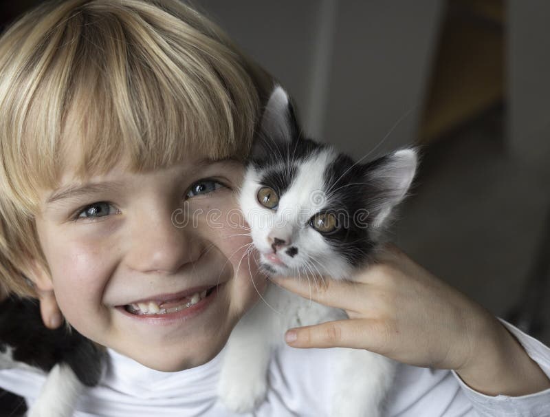 joyful boy smiles with a toothless smile and holds a little beloved black and white kitten near his face