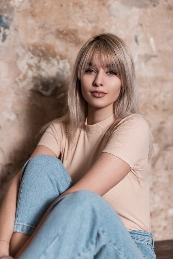 Joyful young amazing woman in a fashionable shirt in vintage blue jeans sit near an old brick wall indoors. Pretty happy girl model enjoys a rest in the studio. Trendy womens wear. Spring style. Joyful young amazing woman in a fashionable shirt in vintage blue jeans sit near an old brick wall indoors. Pretty happy girl model enjoys a rest in the studio. Trendy womens wear. Spring style