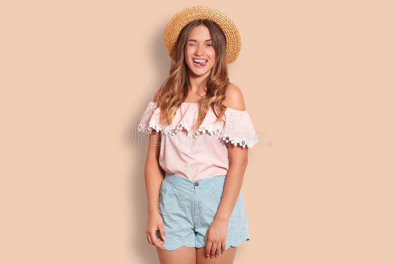 Joyful pretty female with happy expression, blinks eye, shows tongue, wears straw hat, dressed in summer clothing, isolated over s