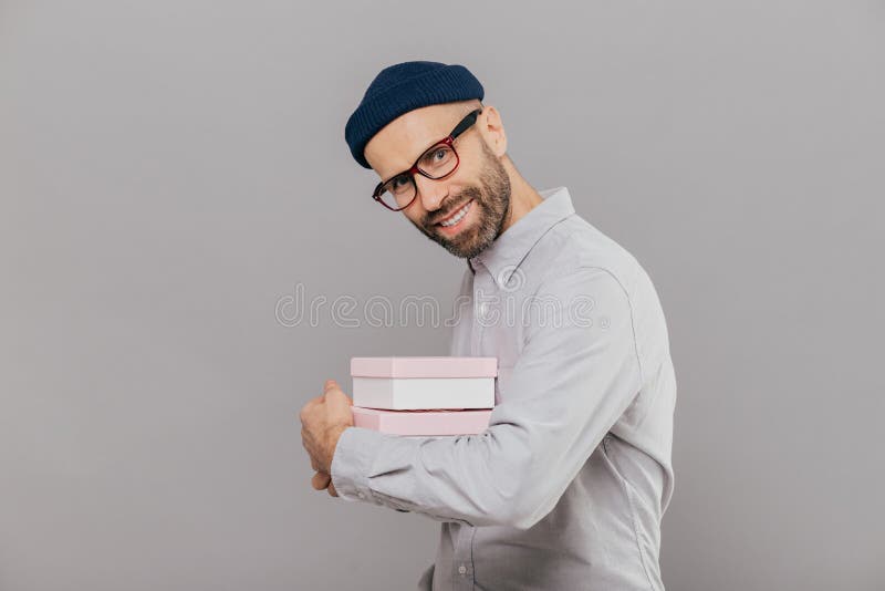 Joyful smiling bearded man with happy expression, dark bristle, wears white shirt and spectacles, carries presents, stands royalty free stock images Joyful smiling bearded man with happy expression, dark bristle, wears white shirt and spectacles, carries presents, stands royalty free stock images