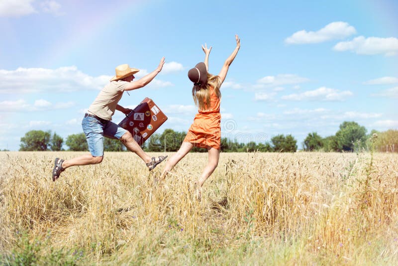 Joyful young couple having fun in wheat field. Excited man and woman running with retro leather suitcase on blue sky