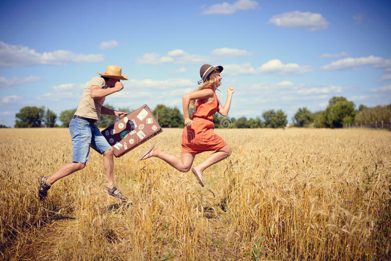 Joyful young couple having fun in wheat field. Excited man and woman running with retro leather suitcase on blue sky