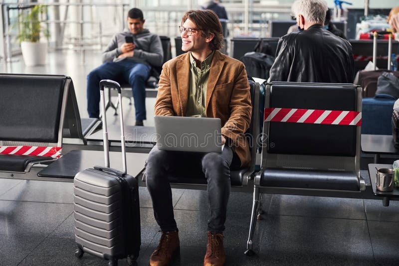 Cheerful male person with notebook on his laps sitting on chair and smiling while waiting for the flight at airport.