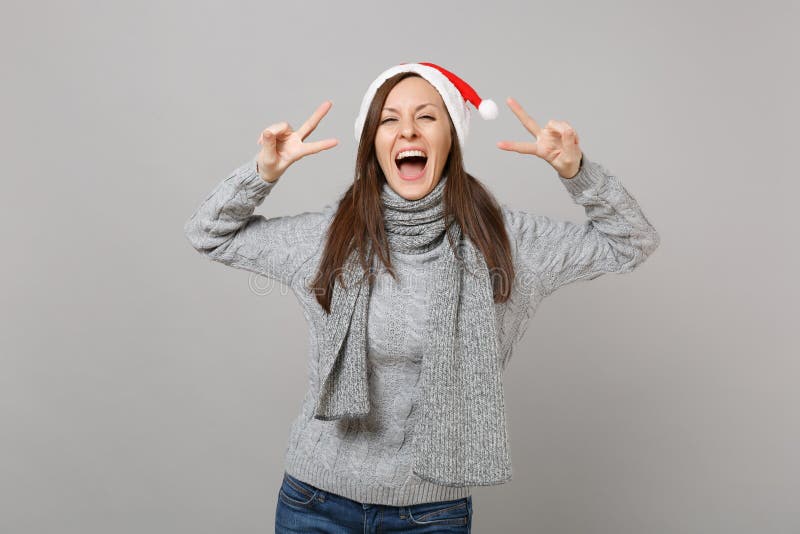 Joyful young Santa girl in gray sweater scarf Christmas hat showing victory sign isolated on grey wall background in stock photography Joyful young Santa girl in gray sweater scarf Christmas hat showing victory sign isolated on grey wall background in stock photography