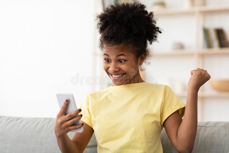 Juego de ganadora de una chica negra en un smartphone sentado en el sofá interior imagen de archivo libre de regalías