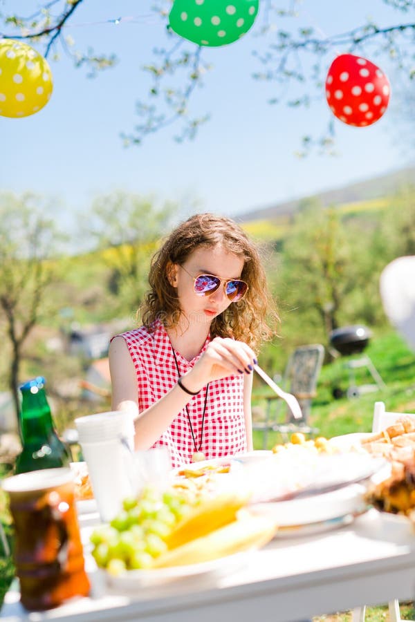 Jugendlich gealtertes Mädchen im roten karierten Hemd, das durch Tabelle auf Gartenfest sitzt lizenzfreies stockbild Jugendlich gealtertes Mädchen im roten karierten Hemd, das durch Tabelle auf Gartenfest sitzt lizenzfreies stockbild