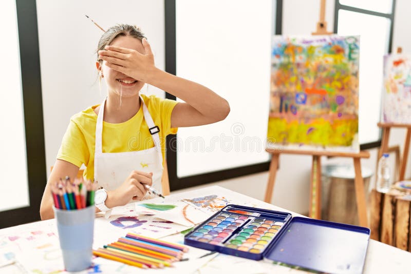 Young brunette teenager at art studio smiling and laughing with hand on face covering eyes for surprise. blind concept. Young brunette teenager at art studio smiling and laughing with hand on face covering eyes for surprise. blind concept
