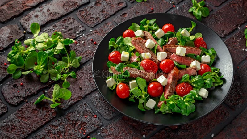 Juicy Beef Sirloin Steak Salad with roasted tomatoes, feta cheese and green vegetables in a black plate. healthy food stock image Juicy Beef Sirloin Steak Salad with roasted tomatoes, feta cheese and green vegetables in a black plate. healthy food stock image